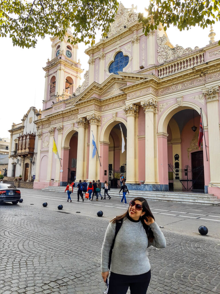 Mujer de frente posando con la Catedral Basílica de Salta de fondo con sus característicos colores pasteles rosas y amarillos. 

Catedral de salta, un sitio que toma su puesto en el listado de lugares sobre qué ver y hacer en Salta