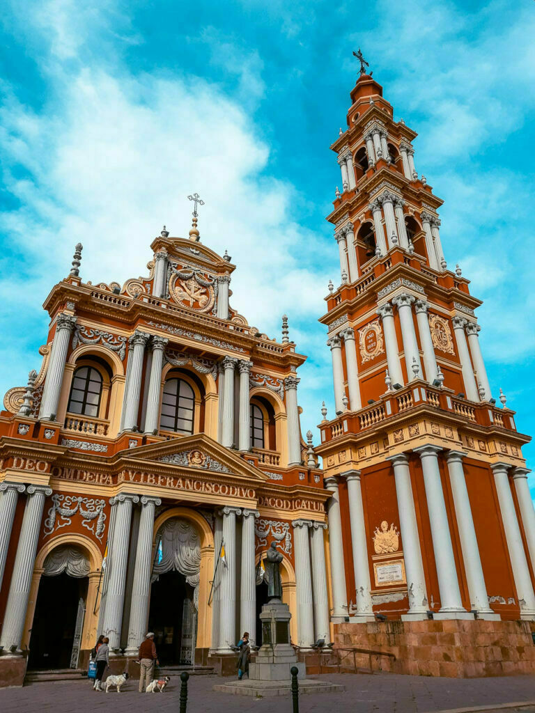Imagen de La Basílica Menor y Convento de San Francisco de la ciudad de Salta, con sus característicos colores rojizos y dorados en un día celeste con nubes que se desparraman a su alrededor