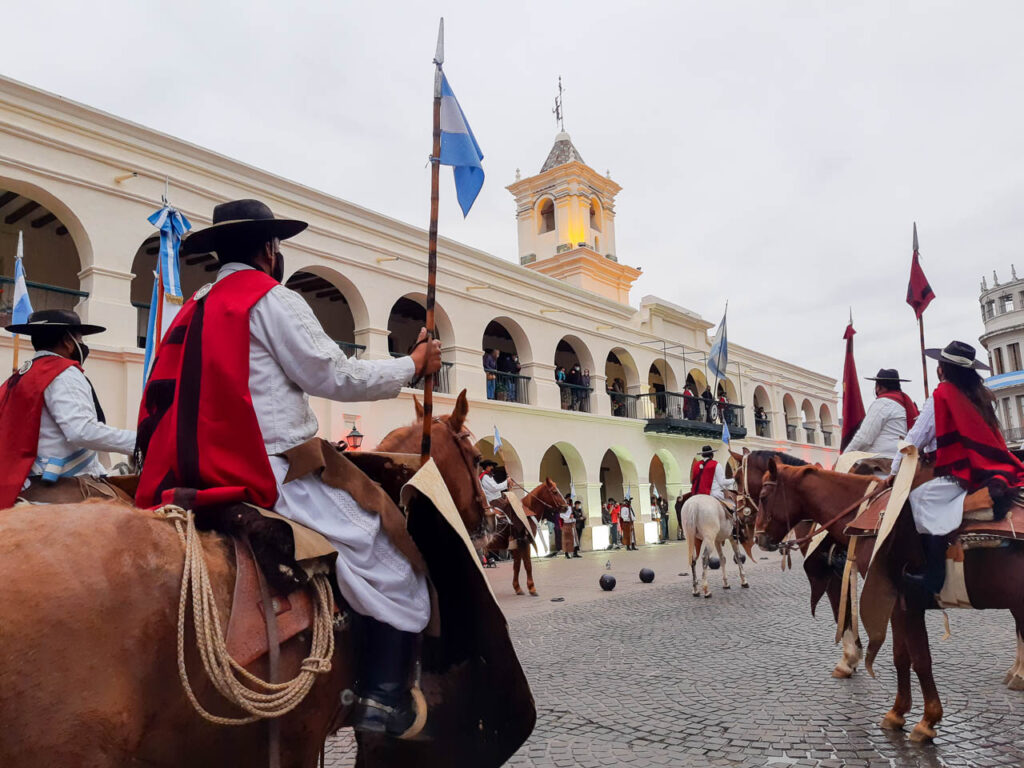 Hombres y mujeres a caballo, vestidos con ropas gauchescas y el característico poncho salteño rojo y negro junto al blanco cabildo de la ciudad de Salta