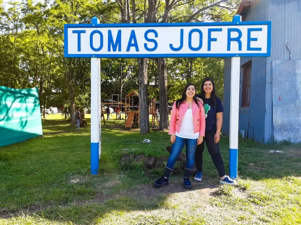 Dos chicas sonriendo a la cámara junto al cartel de la estación de trenes de la localidad de Tomás Jofré
